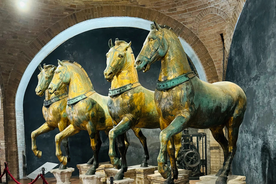 A display of four bronze horse statues in a museum setting with an arched brick background.