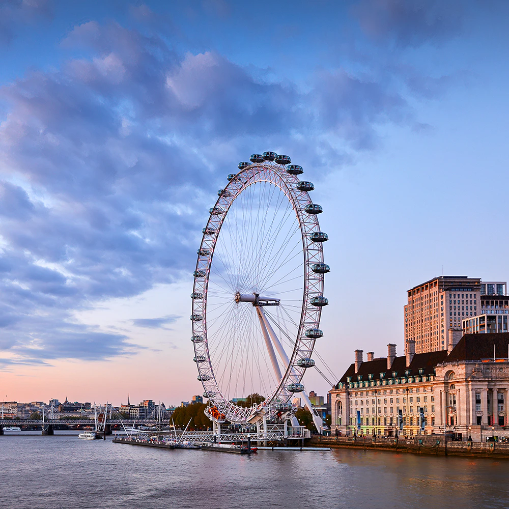 A large Ferris wheel by a riverside with nearby buildings and a bridge under a partly cloudy sky at sunset.