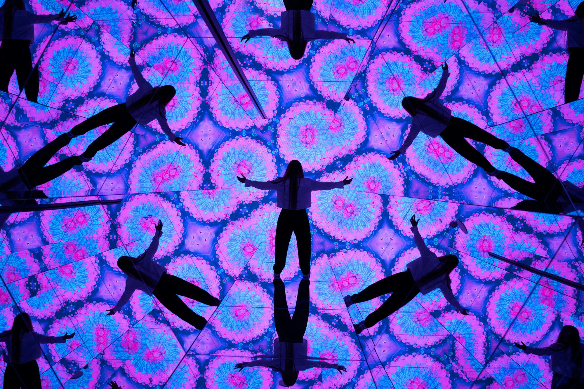 Sylwetka stojąca z wyciągniętymi ramionami, otoczona kalejdoskopowymi odbiciami i żywymi, geometrycznymi wzorami.
