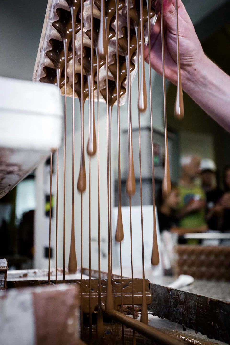 Hand pouring liquid chocolate from a mold, creating long drips into a white tray, with blurred people in the background.