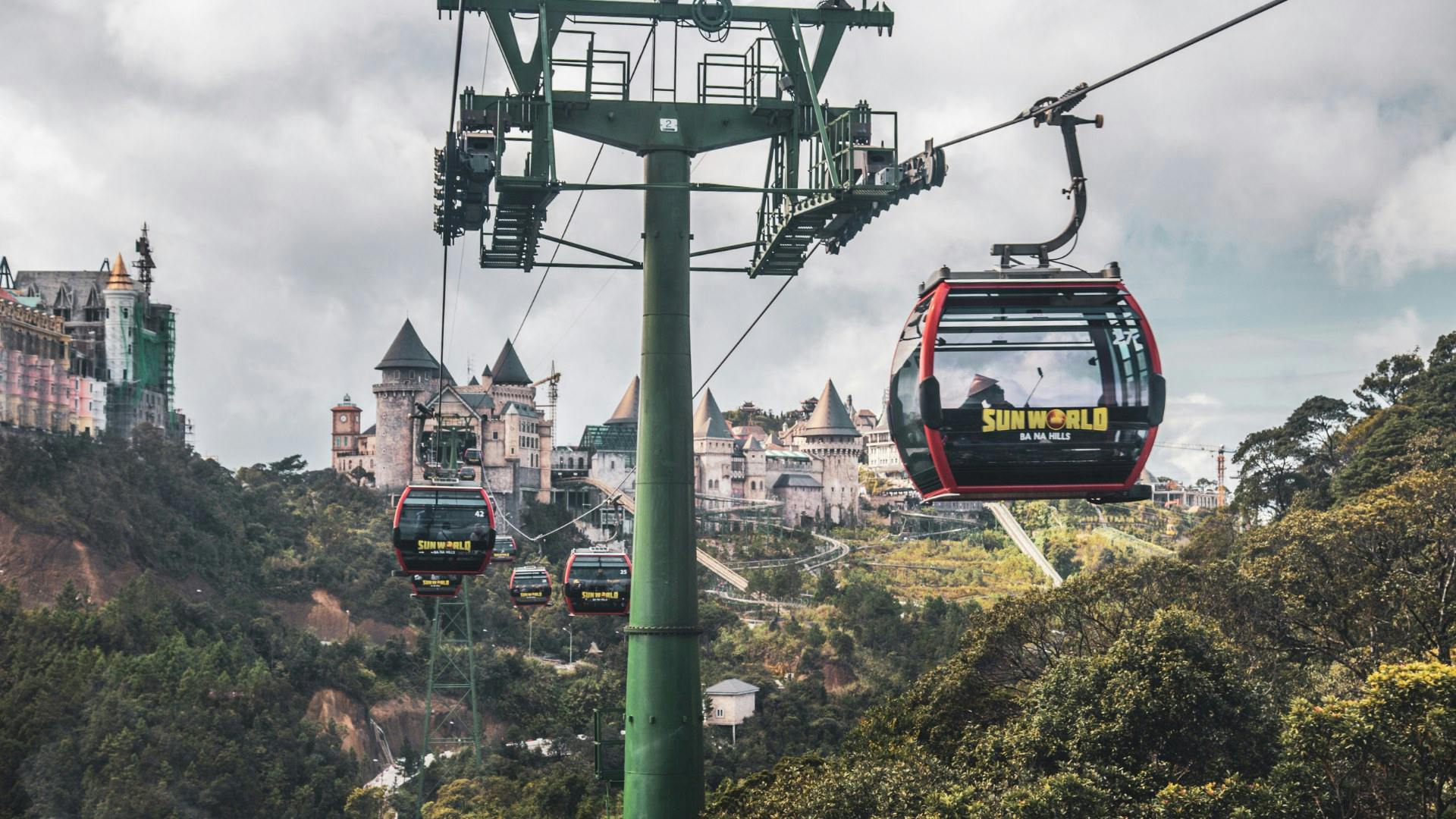 Cable cars with "Sun World" branding travel above lush greenery, with a castle-like structure in the background.