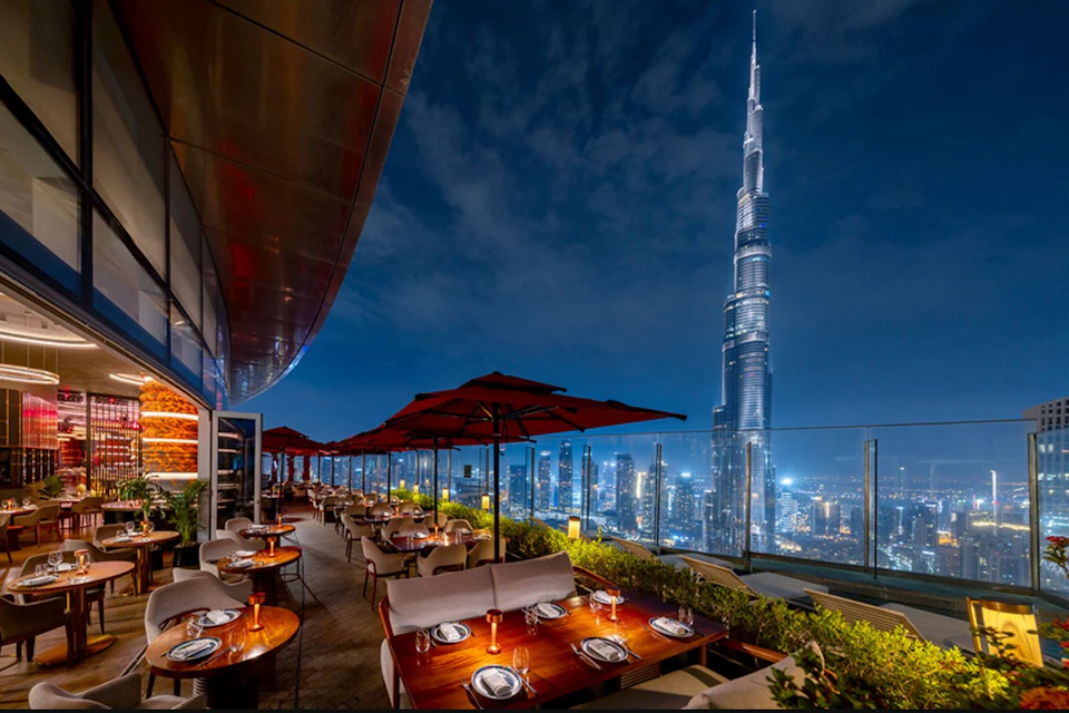Rooftop restaurant with outdoor seating and red umbrellas overlooking a city skyline at night, featuring a tall illuminated skyscraper.