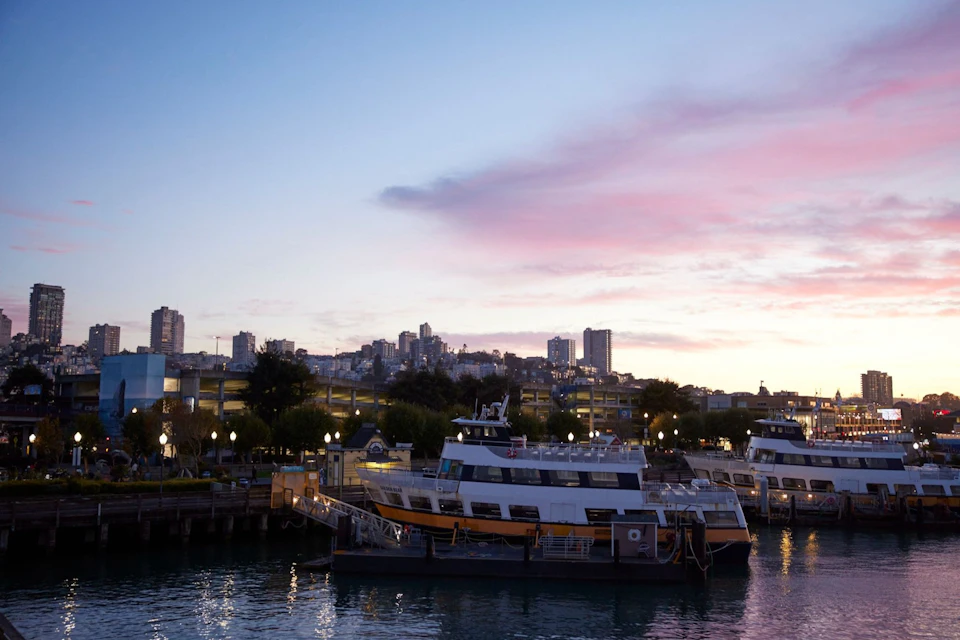 A dock with boats near a city skyline at sunset, with colorful clouds and buildings in the background.