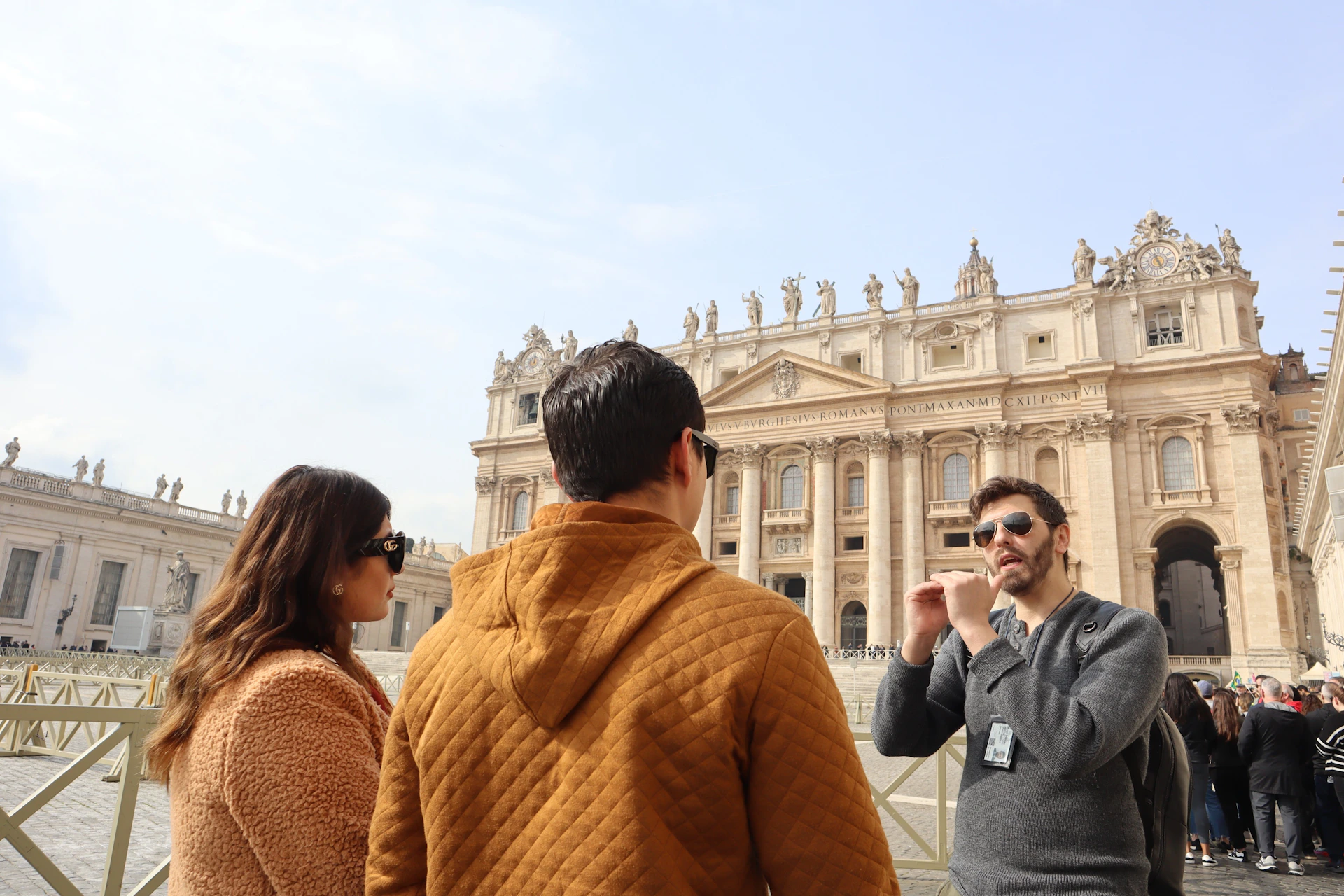 Two people conversing in front of an ornate historical building with sculptures on its roof under a clear sky.