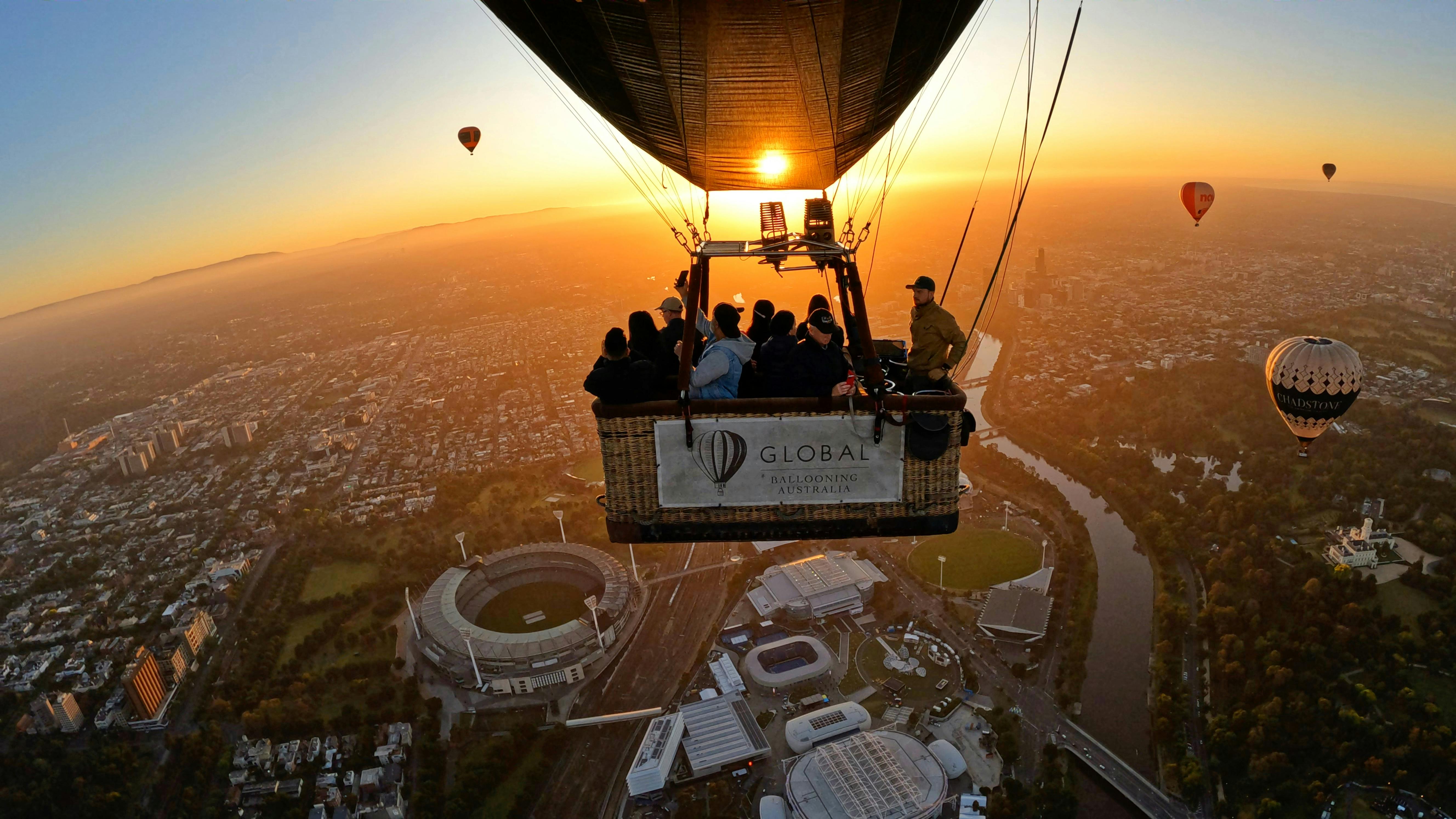 人们乘坐热气球在城市景观上空，那里有体育场馆、河流和另一个日出时可见的气球。