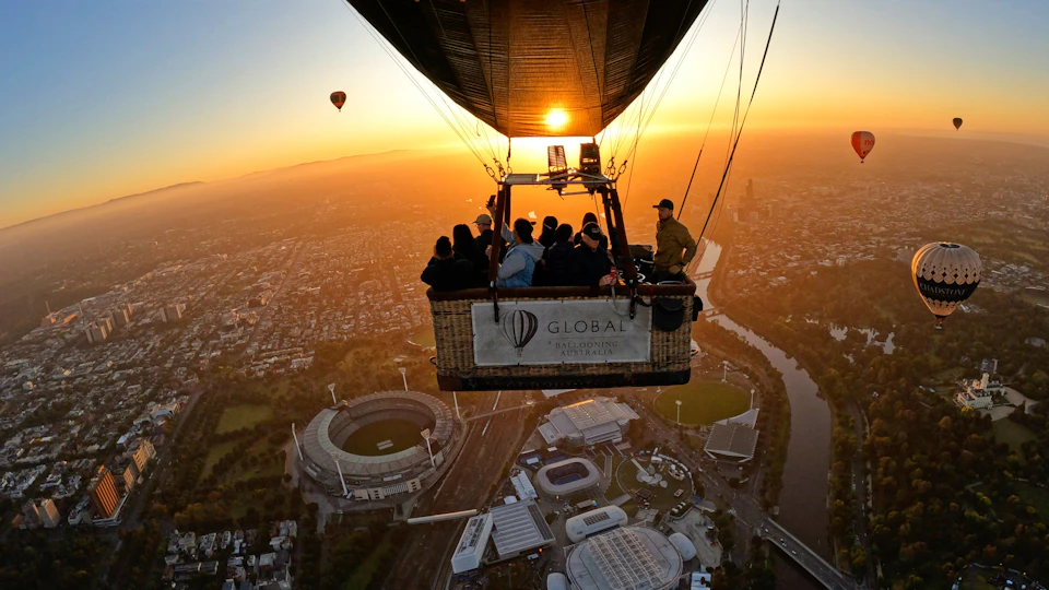 People in a hot air balloon above a cityscape with stadiums, a river, and another balloon visible at sunrise.