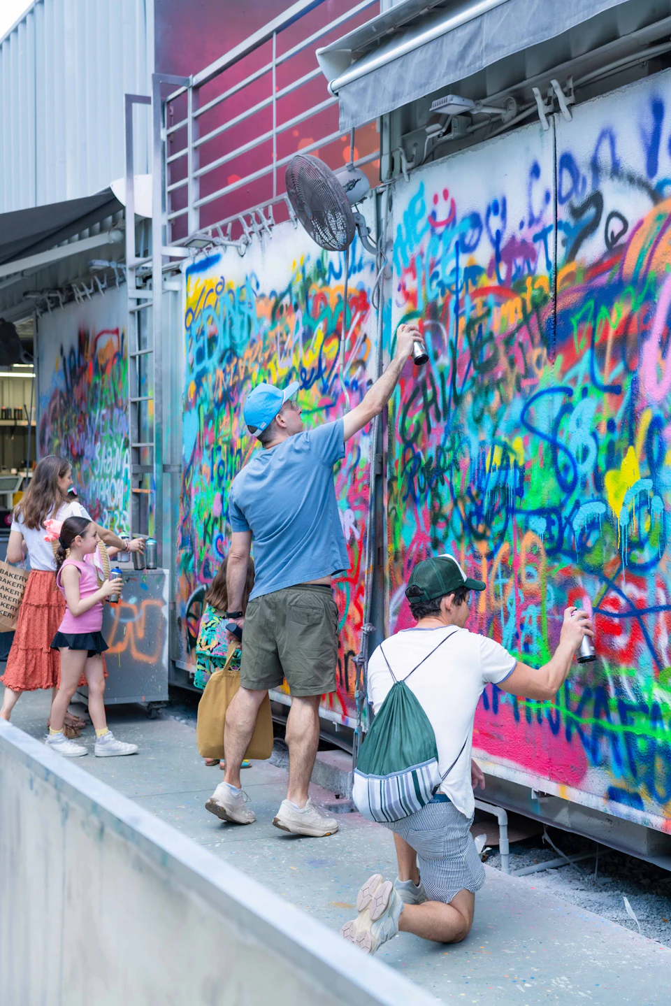 People spray-painting a vibrant graffiti wall with colorful layers of overlapping designs. Some children are also participating.