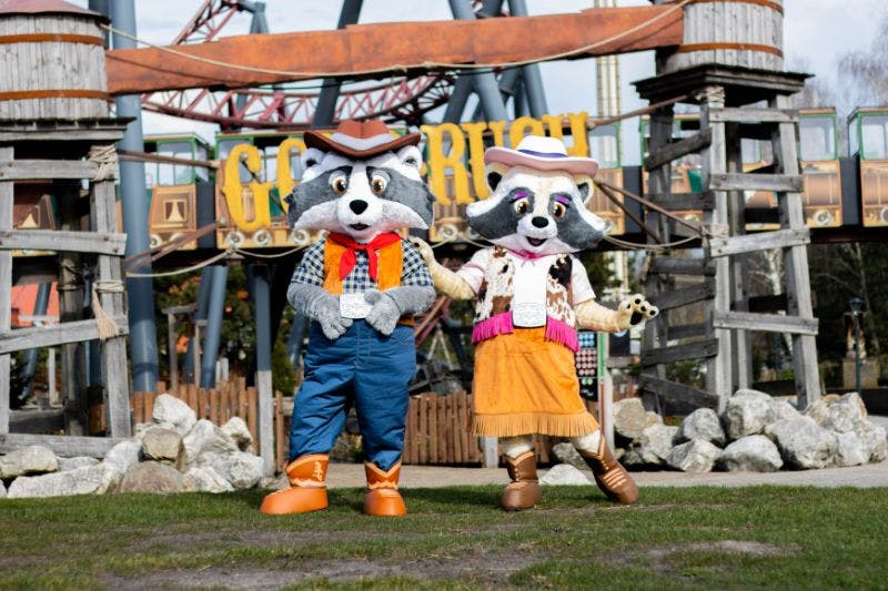 Two raccoon mascots dressed in western attire stand in front of a theme park entrance with the sign "GOLDRUSH."