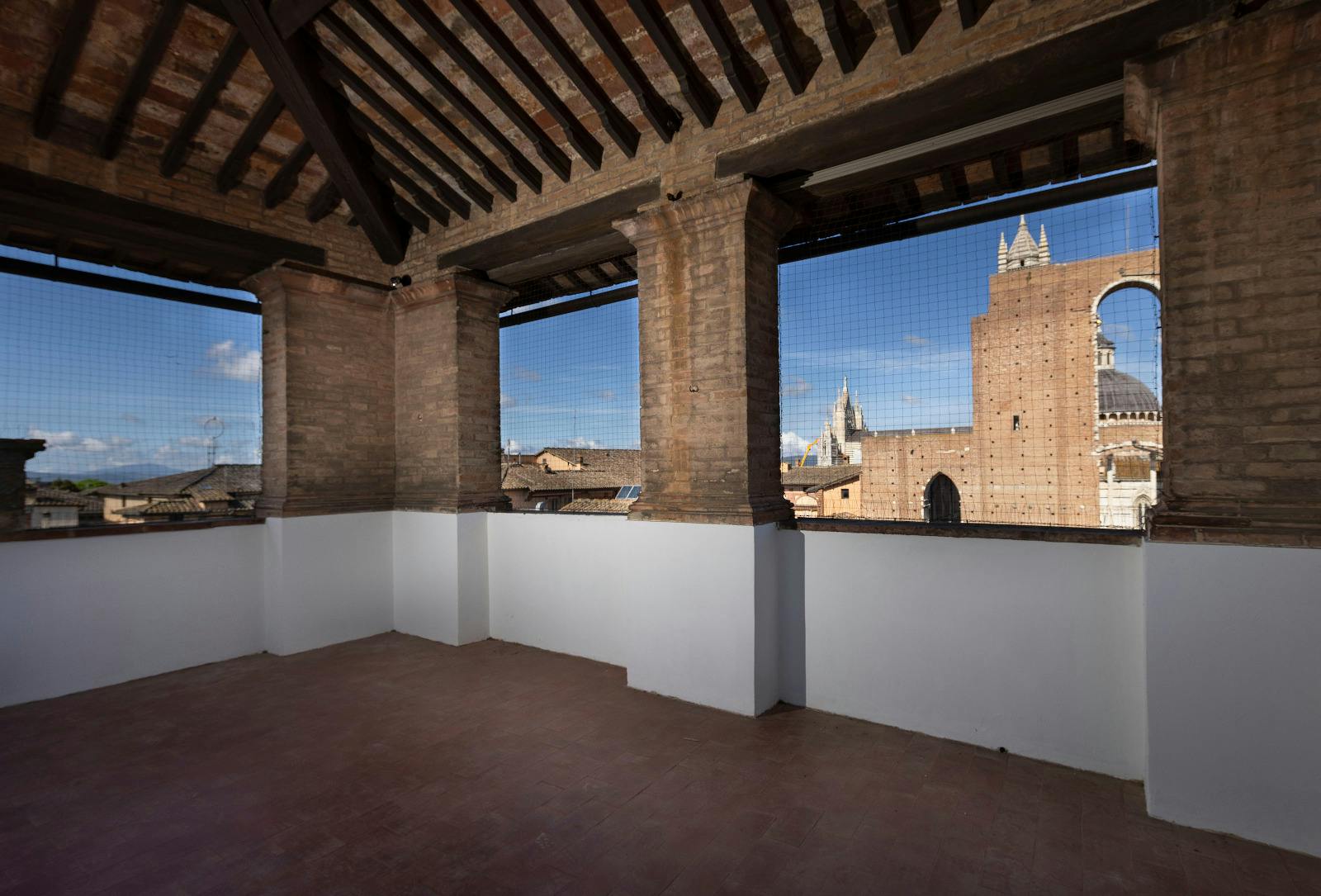 Terrazza aperta con colonne in mattoni e pavimento piastrellato, con vista sulla cattedrale e sugli edifici in lontananza sotto un cielo limpido.