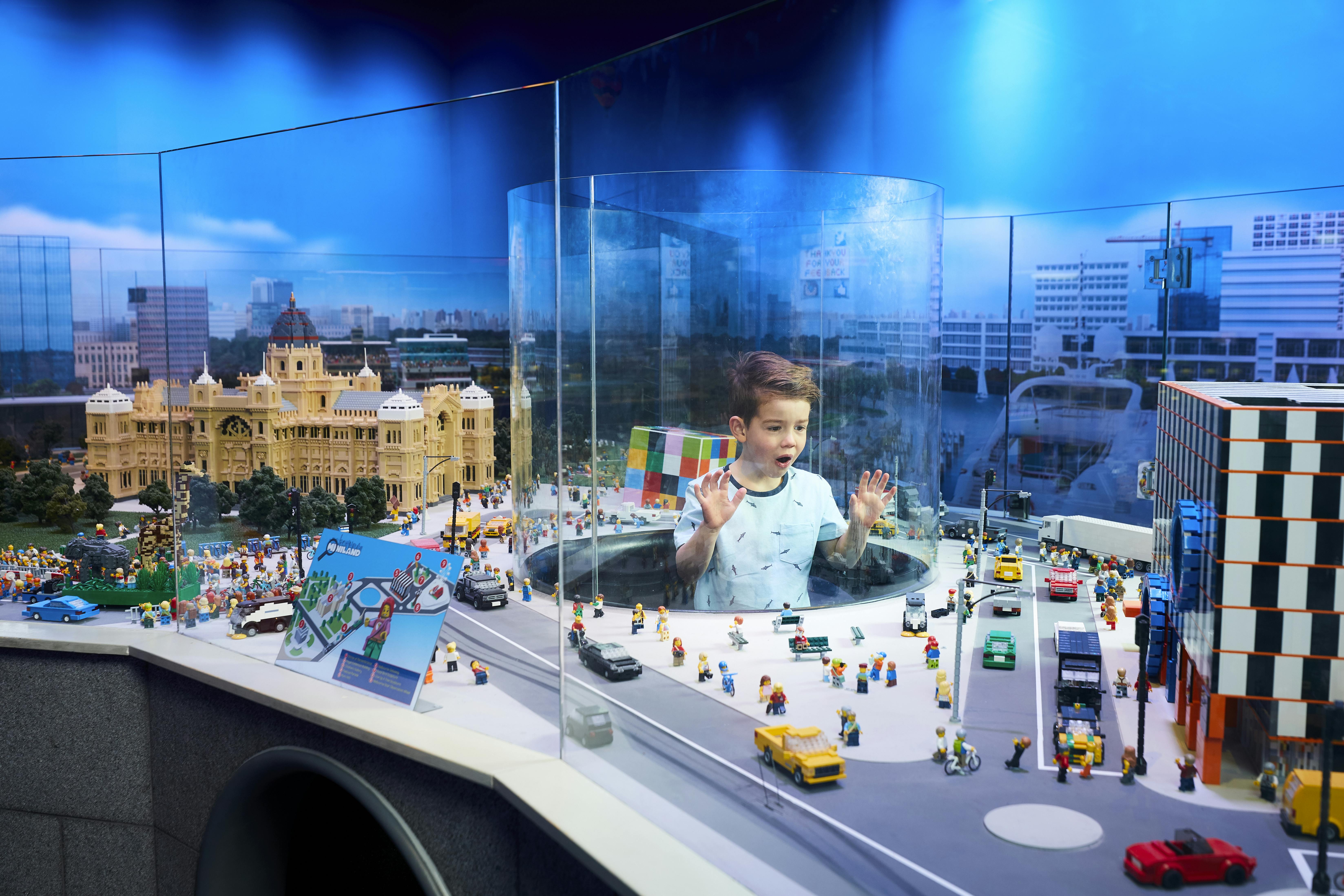 A boy is inside a transparent cylinder in a Lego city display, surrounded by buildings, vehicles, and Lego figures.