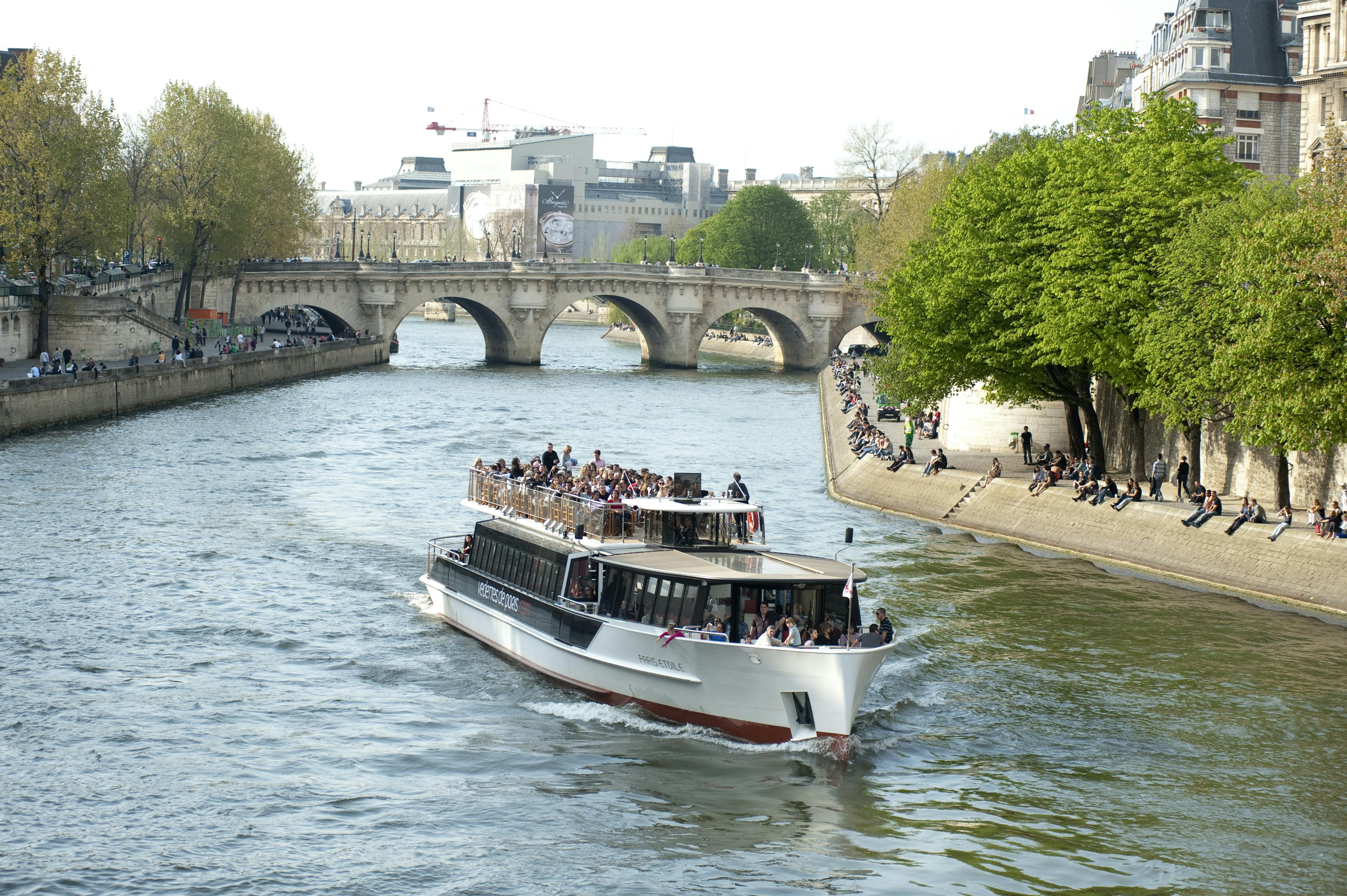 Prohlídkový člun plný lidí se plaví po řece poblíž klenutého mostu s chodci a stromy podél břehů řeky.