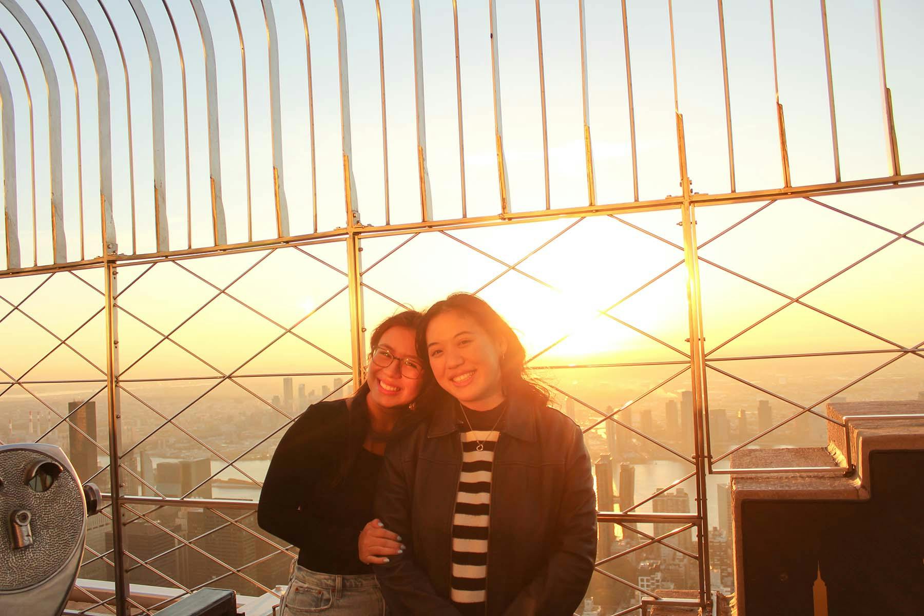 Due donne sorridono e posano insieme su una terrazza panoramica durante il tramonto, con un paesaggio urbano sullo sfondo.