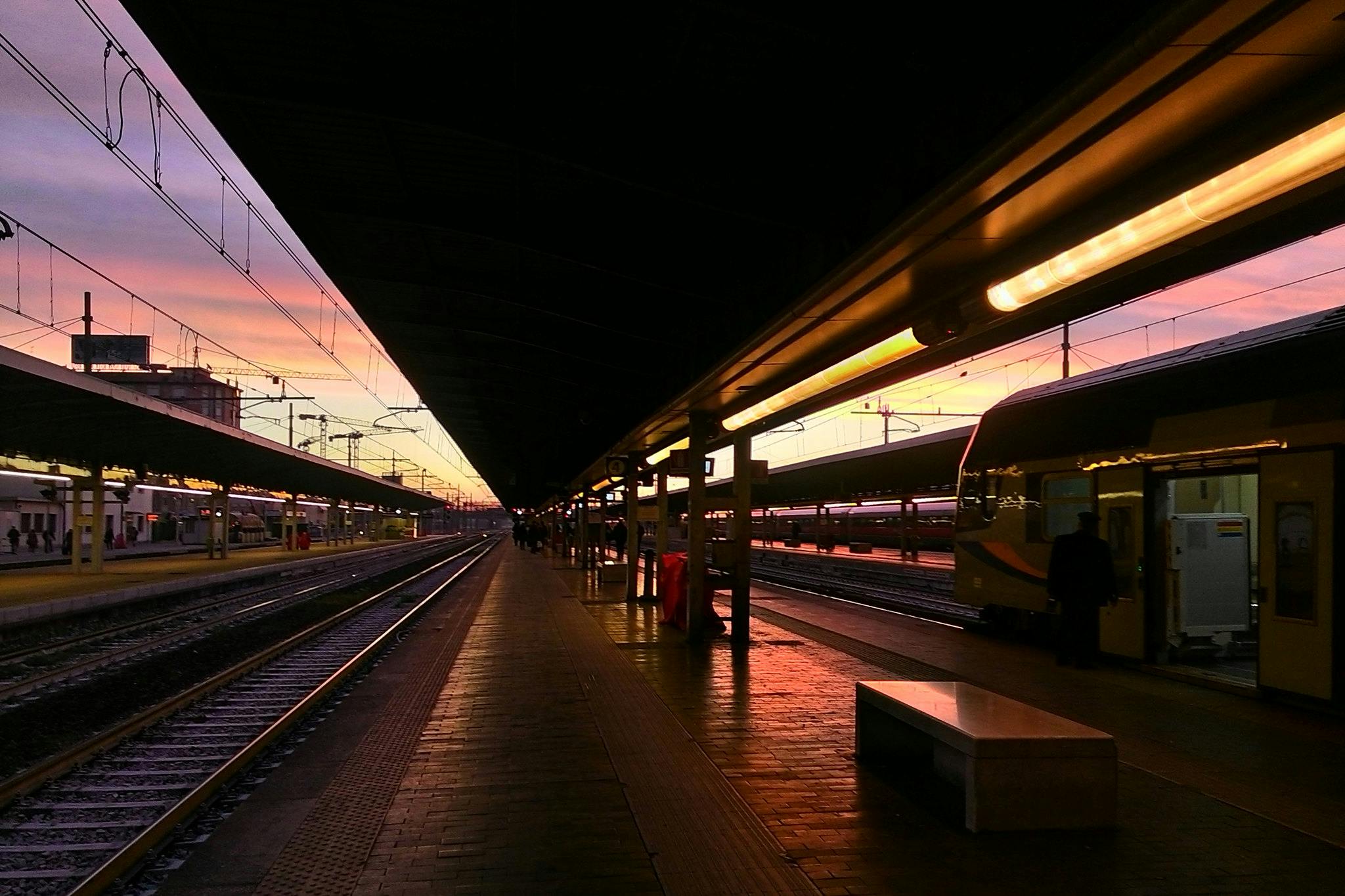 Una plataforma de l'estació de tren al capvespre amb vies a banda i banda i gent pujant al tren. El cel és una barreja de porpra i rosa.