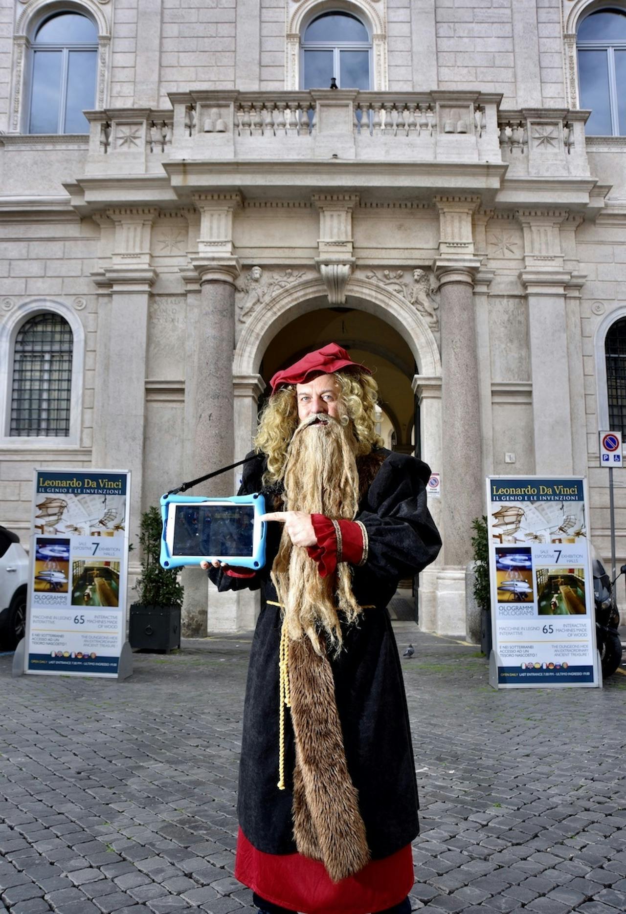 Person in a Leonardo da Vinci costume holding a tablet in front of a historic building with "Leonardo Da Vinci" posters.