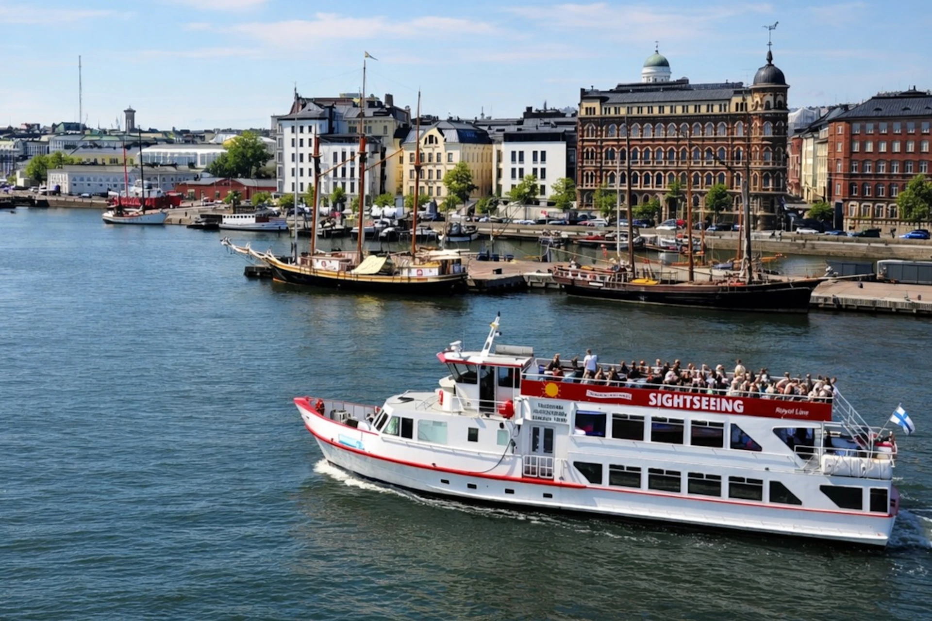 A sightseeing boat on a river with historic buildings and moored ships in the background on a sunny day.