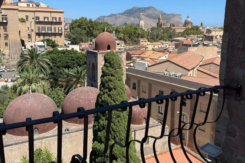 View from a balcony overlooking a city with red-domed buildings, lush greenery, and distant mountains under a clear blue sky.