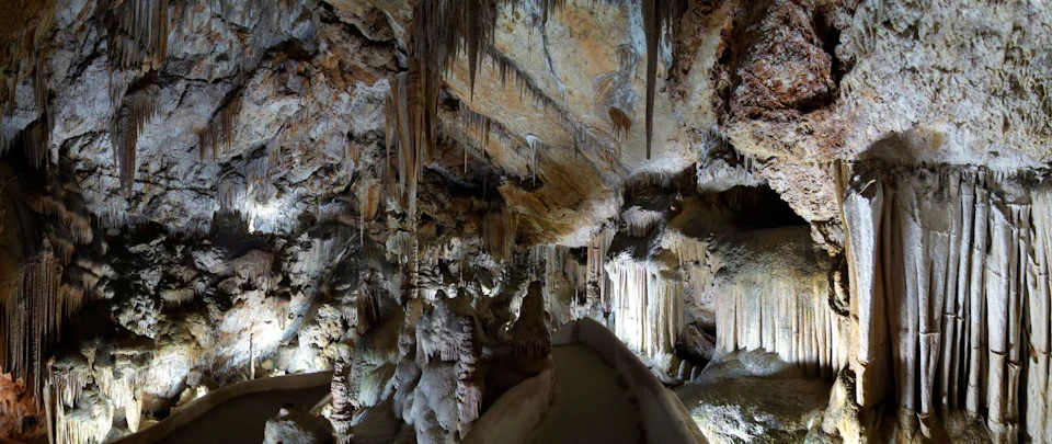 A cave interior filled with intricate stalactites and stalagmites along a narrow, illuminated pathway.