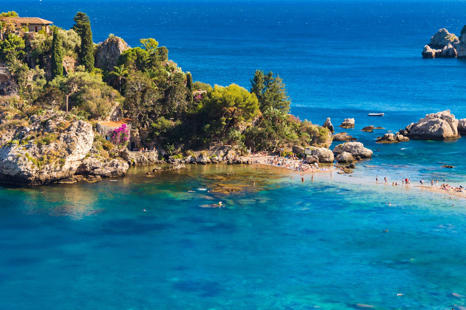 Small beach with people sunbathing and swimming, surrounded by rocky cliffs and lush greenery, near a vibrant turquoise sea.