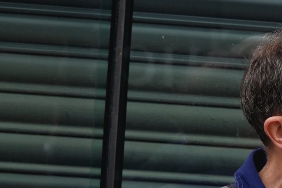 Two men are talking outside next to a building with closed metal shutters. One man holds a crinkled brown paper bag.