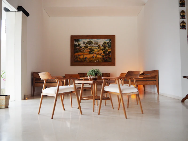 A minimalist dining room with a wooden table, chairs, a potted plant centerpiece, and a framed landscape painting on the wall.
