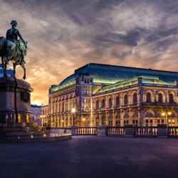 Ryttarstaty på ett torg i skymningen, med en upplyst historisk byggnad och dramatisk himmel i bakgrunden.