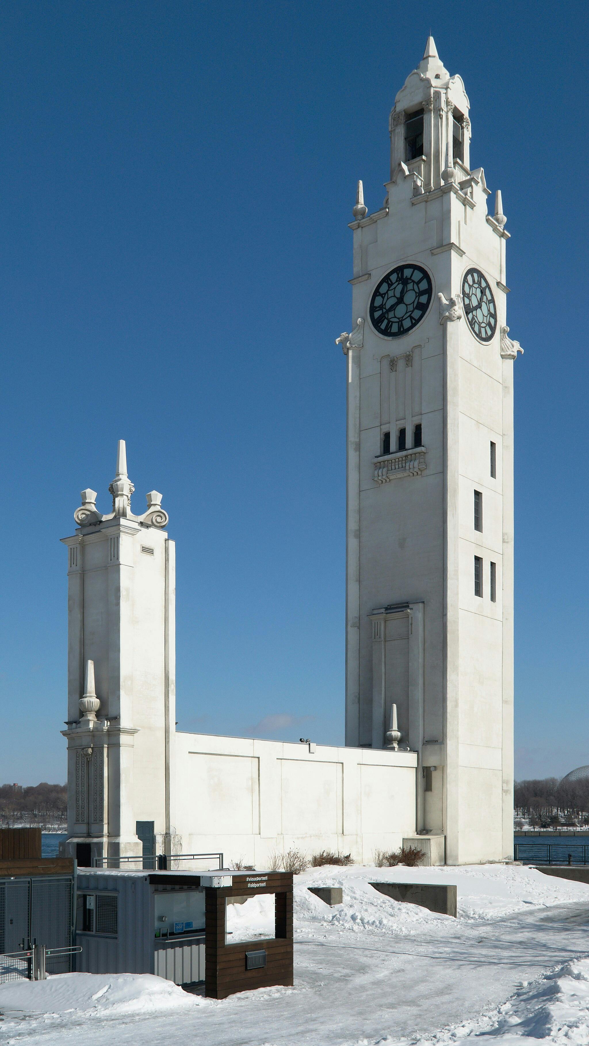 Tall white clock tower with ornate details stands beside a smaller structure, set against a bright blue sky and snowy landscape.