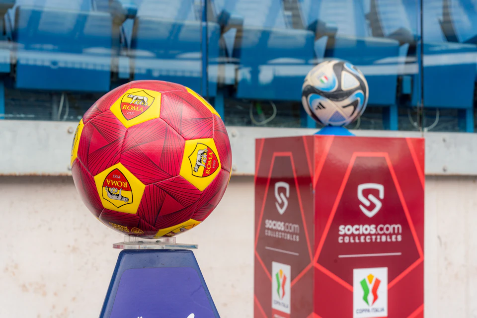 Two soccer balls on display stands, one red with Roma logos and the other multicolored, near branded panels in a stadium.