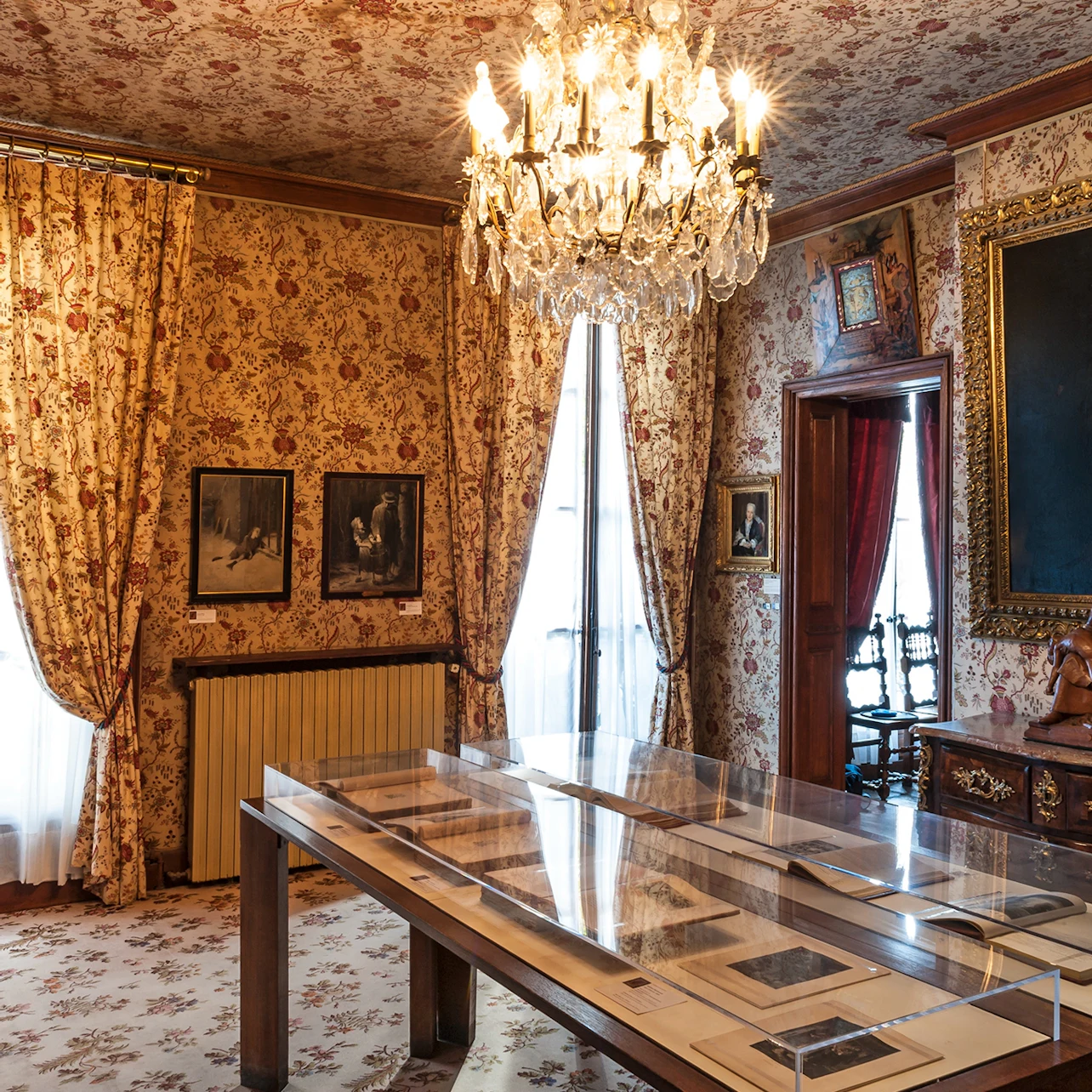 Room with floral wallpaper, chandelier, and framed artwork. Table with display cases in the center, and ornate wooden furniture.