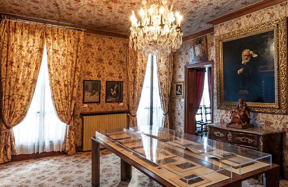 Room with floral wallpaper, chandelier, and framed artwork. Table with display cases in the center, and ornate wooden furniture.