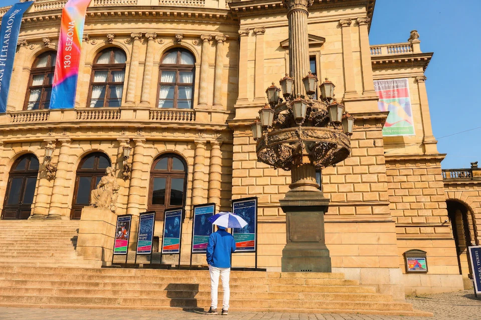 A person with a blue jacket and umbrella stands in front of a historic building with posters displayed on stands.