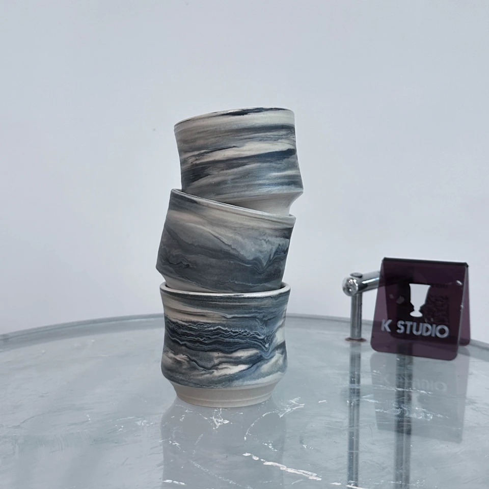 Three ceramic cups with a marbled black and white design stacked on a clear surface. A sign with "K Studio" is nearby.