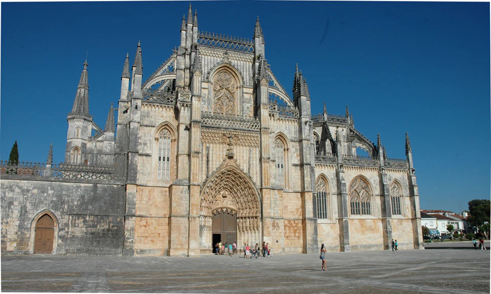 A large, intricately detailed Gothic cathedral with people gathered at its entrance and a person taking a photo in front.