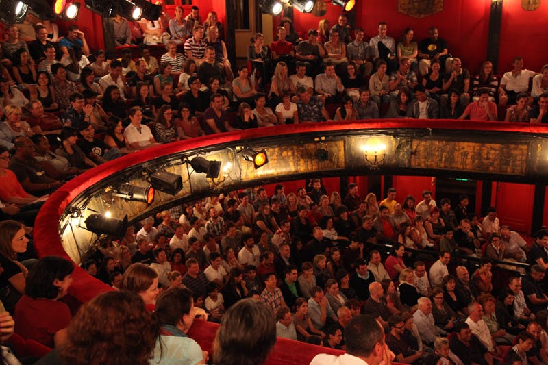 一大群观众坐在一个红色座位的圆形剧院里，被多个舞台灯光照亮，所有人都专注于一场看不见的表演。