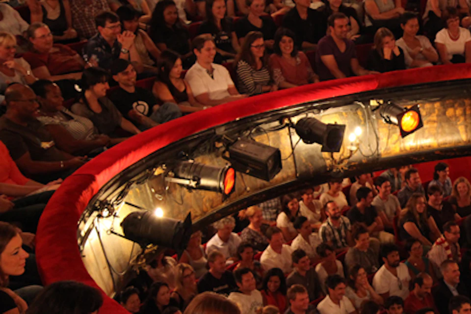 A large audience sits in a round theater with red seating, lit by multiple stage lights, all focused on an unseen performance.