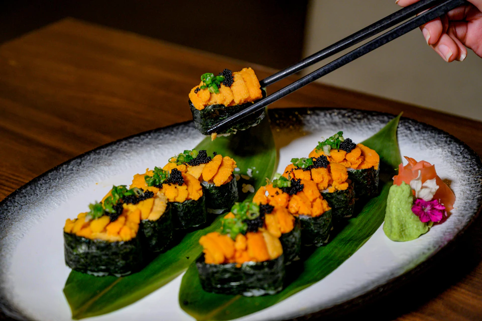 A plate of sushi garnished with sea urchin, green onions, and roe, presented on green leaves with chopsticks picking one piece.