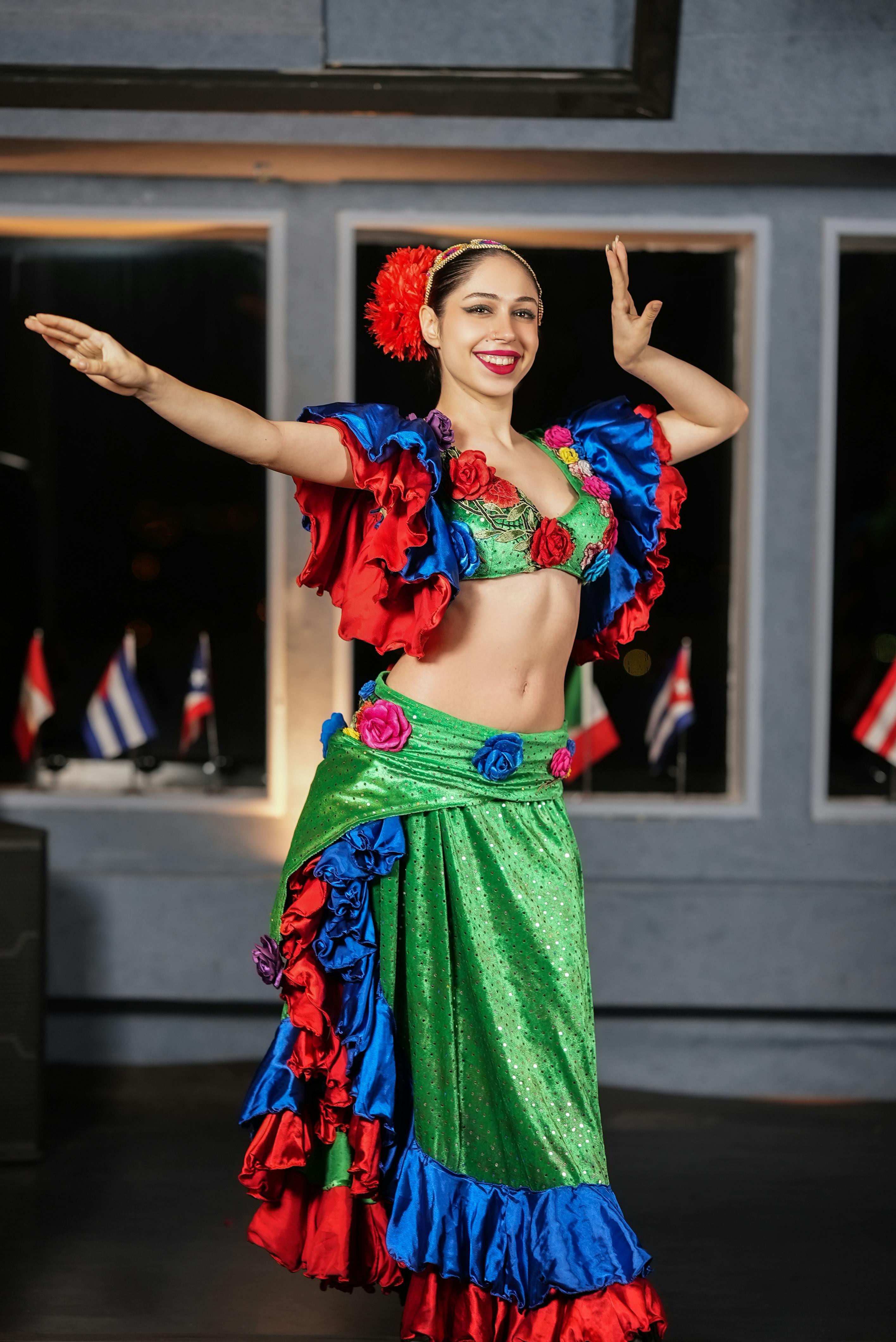 カラフルなフリルのコスチュームを着た笑顔の女性が、腕を広げてポーズをとっています。背景には旗が表示されています。