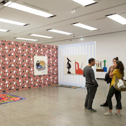 Tre personer samtalar i ett modernt konstgalleri med färgglada mönstrade väggar och abstrakta målningar.