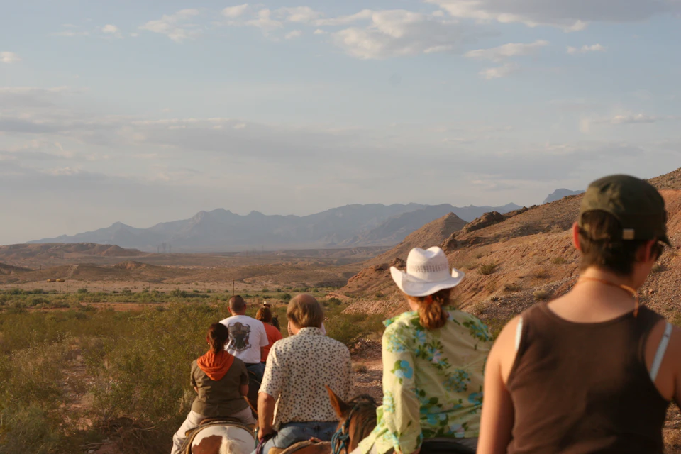 A group of people riding horses on a trail through a desert landscape with mountains in the background under a partly cloudy sky.