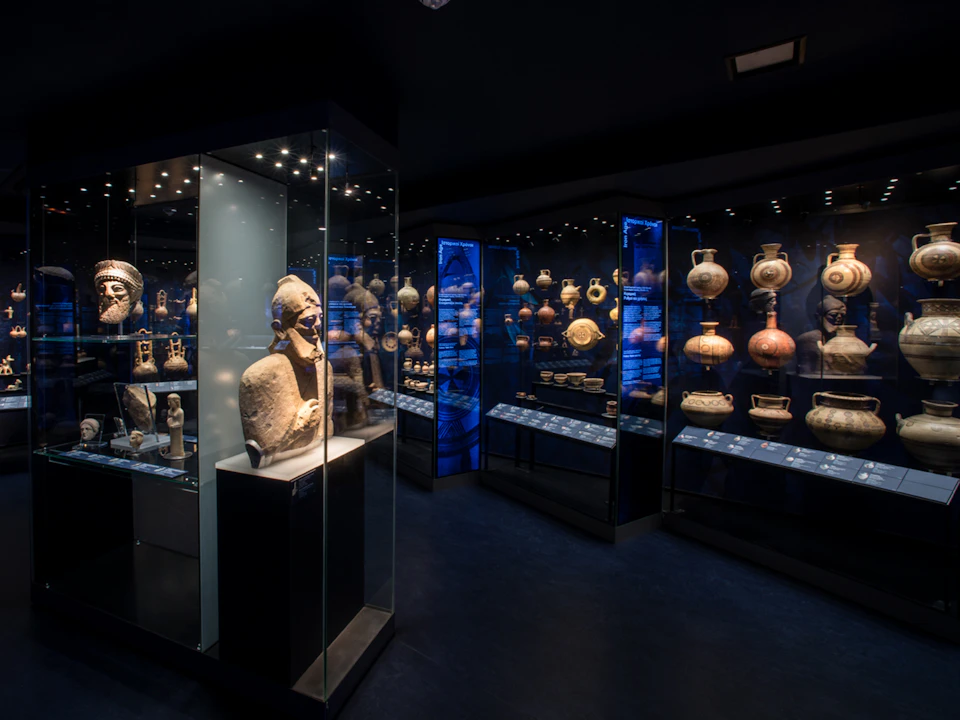 A dimly lit museum exhibit displaying various ancient artifacts and pottery inside glass cases.