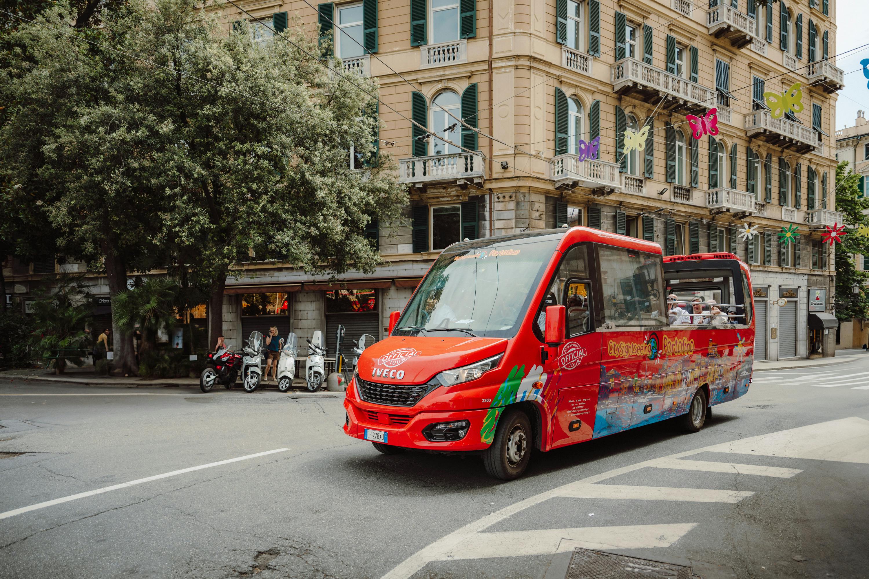 Red tourist bus on a city street with scooters parked near a large building adorned with balconies and colorful butterfly decorations.