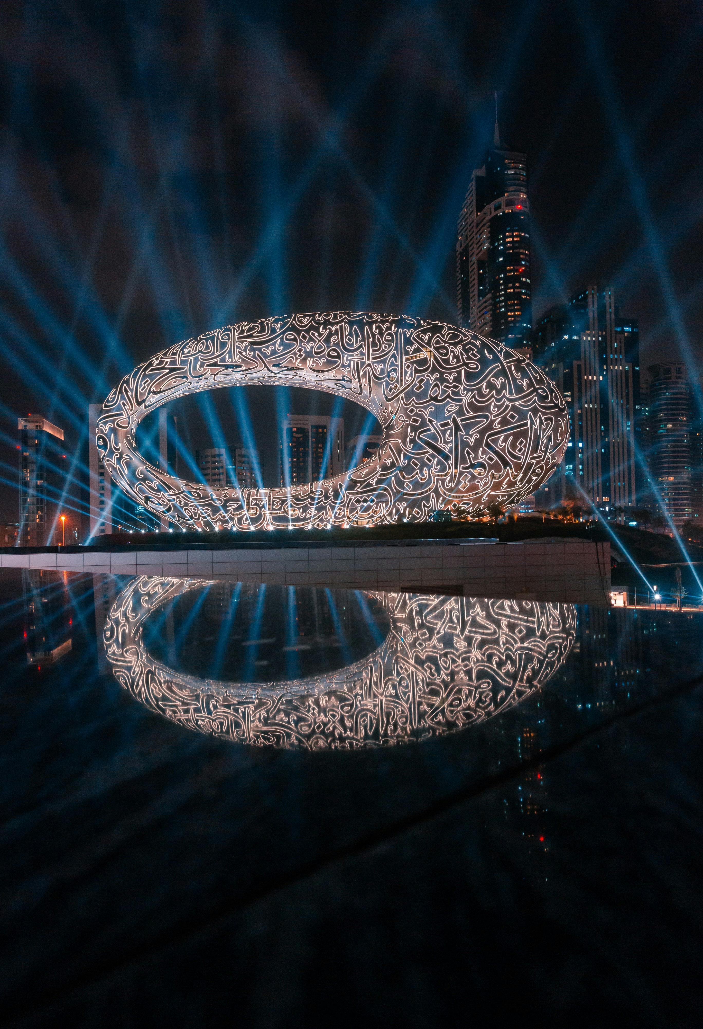 这座未来主义的椭圆形建筑拥有发光的脚本图案，与摩天大楼的城市景观相映成趣，并由蓝色光束照亮。