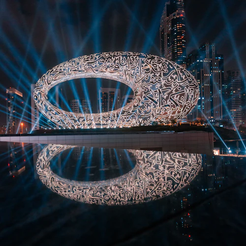 Ein futuristisches, ovales Gebäude mit leuchtenden Schriftmustern vor einer Stadtlandschaft mit Wolkenkratzern, die von blauen Strahlen beleuchtet wird.