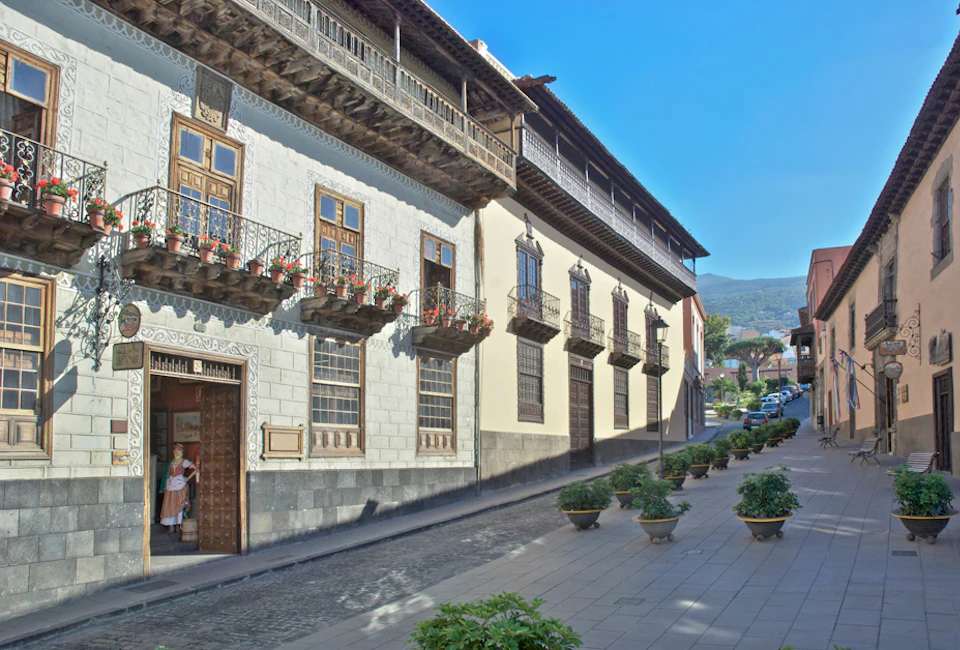 A street with historic buildings featuring wooden balconies and flower pots. Potted plants line the sidewalk under a clear blue sky.