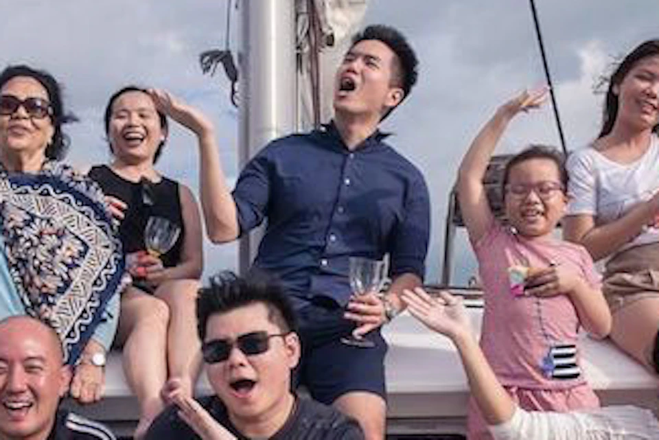 A group of people smiling and posing energetically on a sailboat with a netted seating area, under a partly cloudy sky.