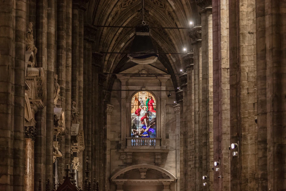 An ornate cathedral interior with a large stained-glass window depicting a colorful biblical scene.