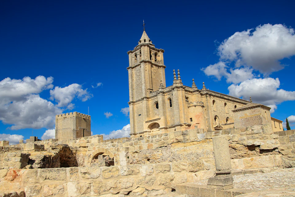 A historical stone church with a tall bell tower, surrounded by ruins under a bright blue sky with few clouds.
