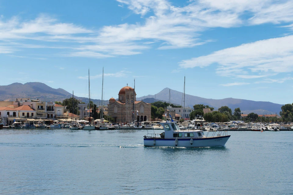 A small fishing boat sailing on a calm waterfront with a town, a church with a dome, and mountains in the background.