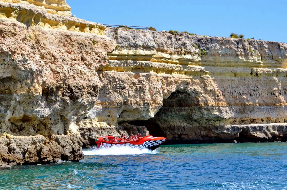 A red speedboat with passengers cruises near rocky cliffs over clear blue water on a sunny day.
