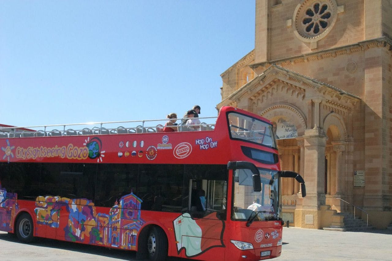 Red double-decker bus with open top passes in front of a historic stone building under a clear blue sky. People are seen on the top deck.