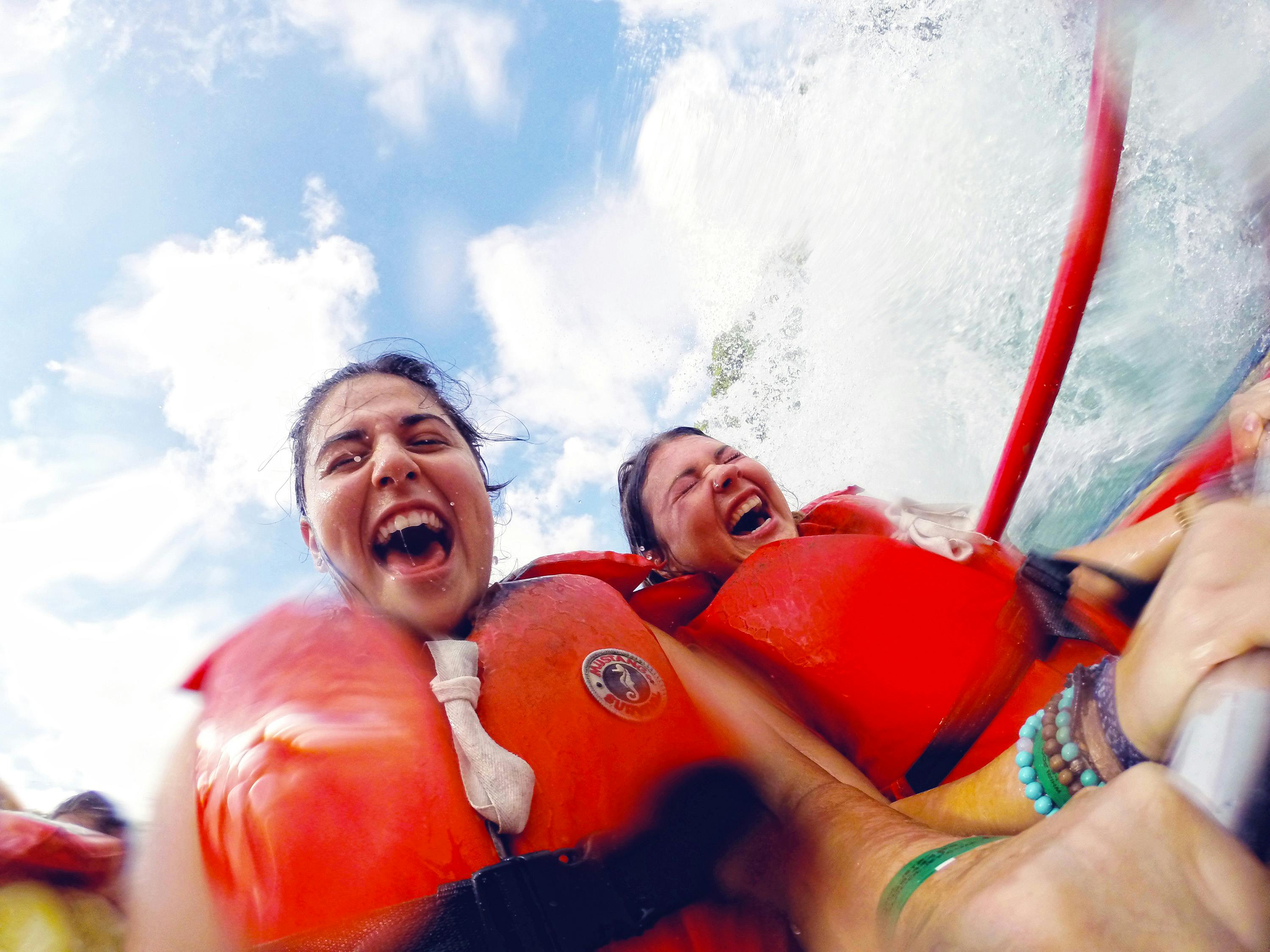 Due persone con giubbotti di salvataggio rossi che ridono e si tengono su una zattera, con spruzzi d'acqua e un cielo azzurro sullo sfondo.